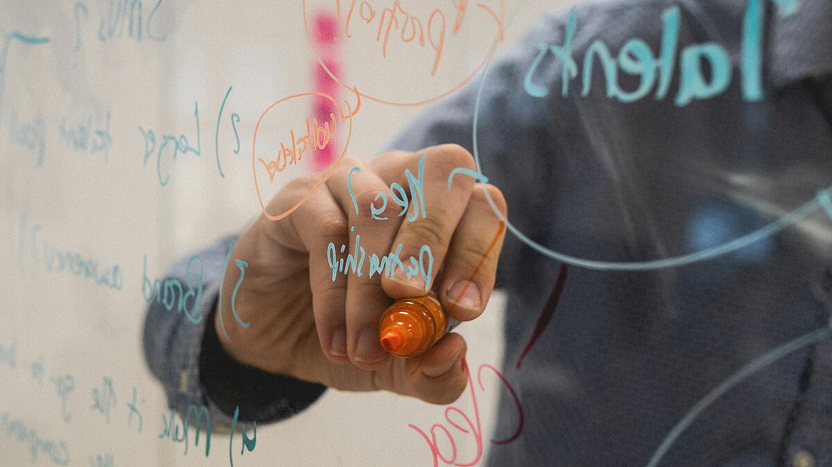 Zu sehen ist eine durchsichtige Schreibtafel, auf der eine Person mit orangem Stift schreibt.