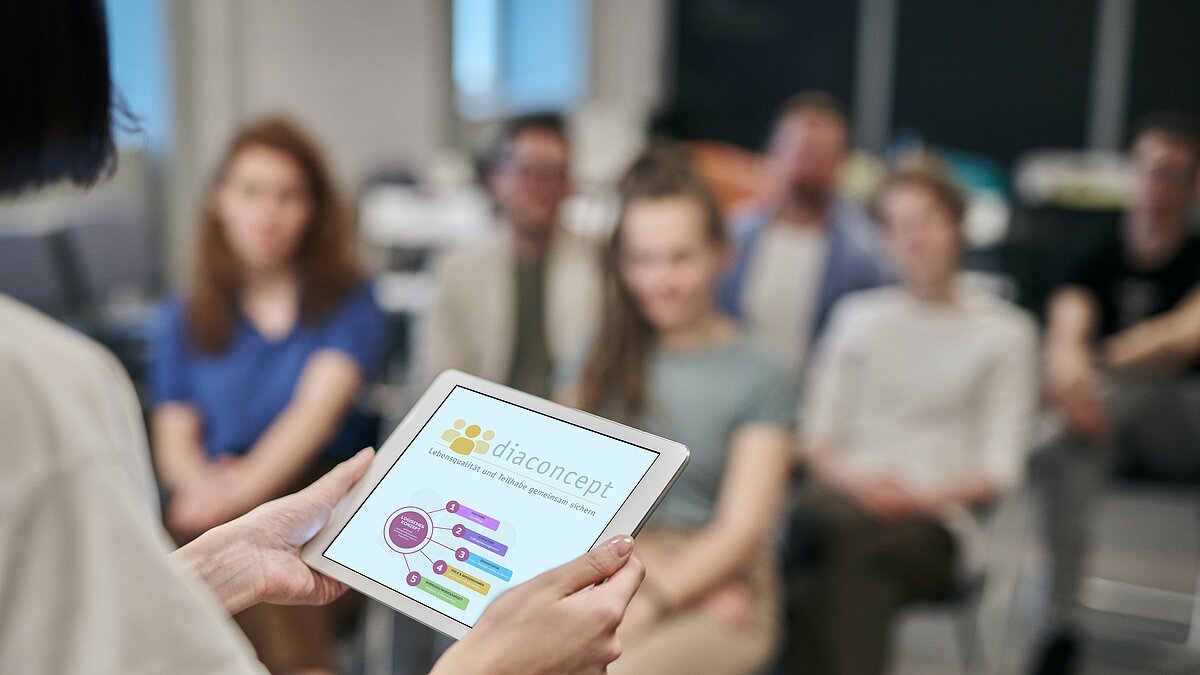 Zu sehen ist eine Referentin, die ein Tablet in den Händen hält und in Richtung von Seminarteilnehmer:innen einen Vortrag hält.