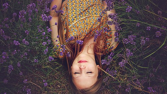 Zu sehen ist eine rothaarige Frau. Sie liegt entspannt in einem Lavendel-Feld.