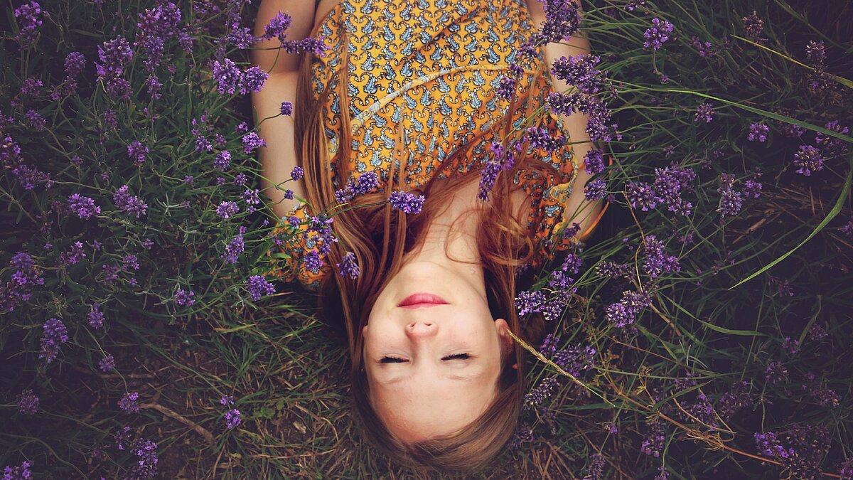 Zu sehen ist eine rothaarige Frau. Sie liegt entspannt in einem Lavendel-Feld.
