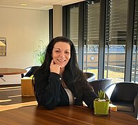 Zu sehen ist eine Frau mit langen dunklen Haaren, die eine dunkle Bluse trägt, die an einem Stehtisch steht und leicht auf ihren rechten Arm gestützt in die Kamera blickt und lächelt. 