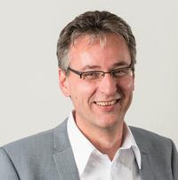 Zu sehen ist ein Mann mit Brille und kurzen dunkelgrauen Haaren, der einen hellgrauen Anzug und ein weißes Hemd trägt und in die Kamera lächelt. 